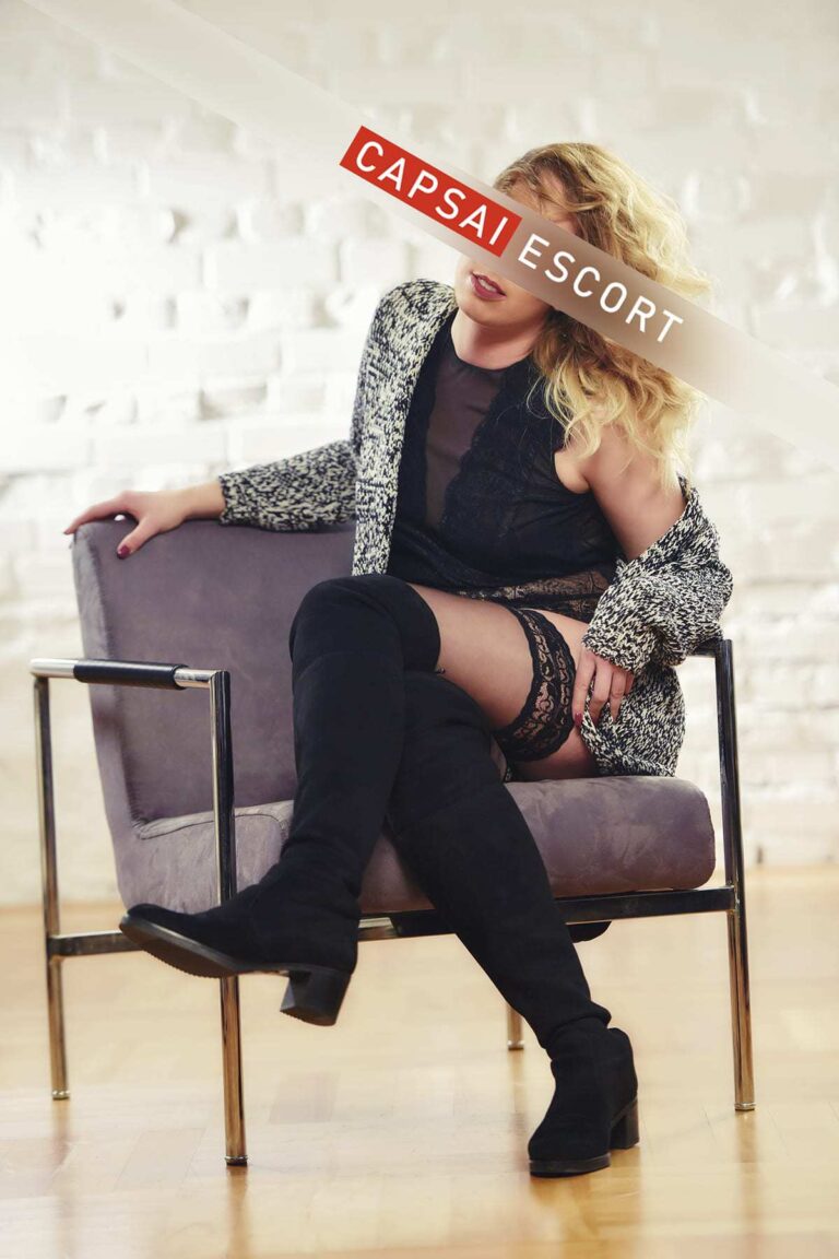 Escort Dame Esra in einem schwarzen Spitzenbody und schwarzen Nylons und schwarzen Overknees in einem Sessel sitzend.