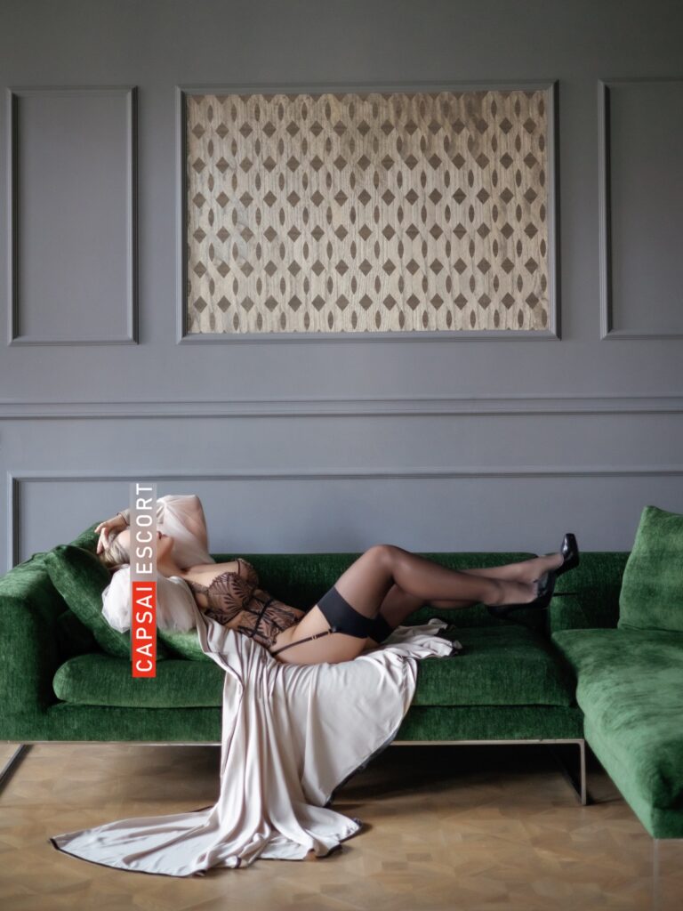 Escort Dame Freya in schwarz/beigen Dessous und schwarzen Nylons mit High Heel und einem langen Morgenmantel auf dem Sofa liegend, mit Ansicht von der Seite.