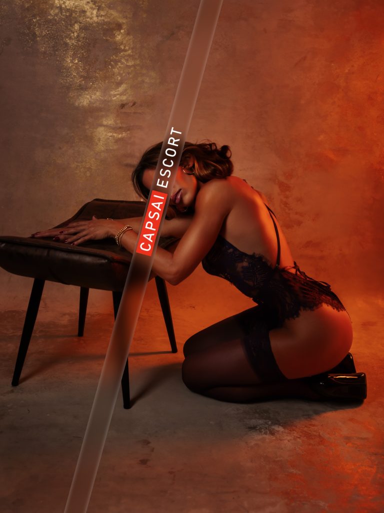 Escort Dame Heidi in einem schwarzen Spitzenbody mit schwarzen Nylons und High Heels in einer kniender Position vor einem Hocker.