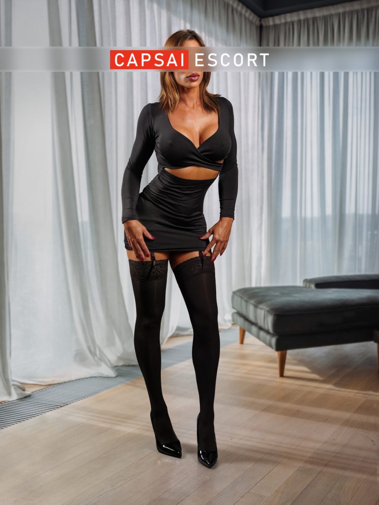 Escort Dame Heidi in einem schwarzen Kleid mit Ausschnitten und schwarzen High Heels mit schwarzen Nylons.