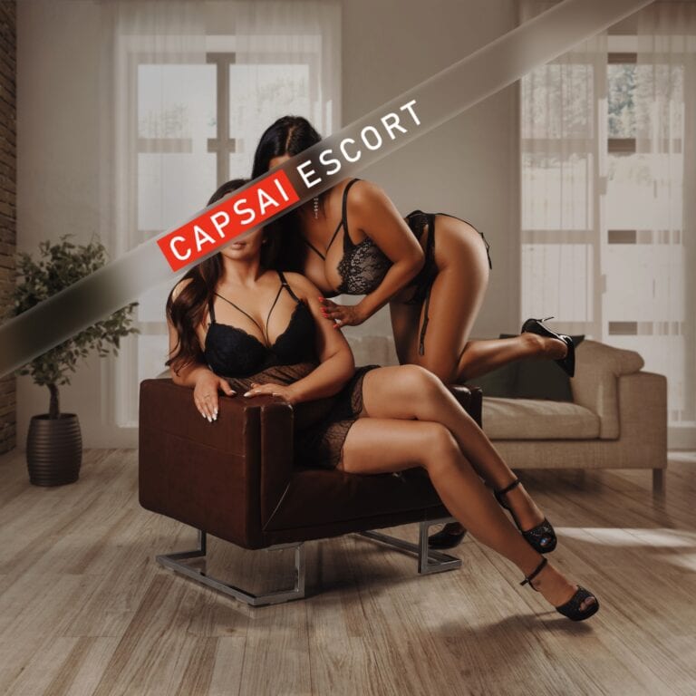 Escort Dame Isabelle in schwarzen Dessous und High Heels im Sessel sitzend, dahinter eine Dame in Dessous auf der Armlehne kniend.