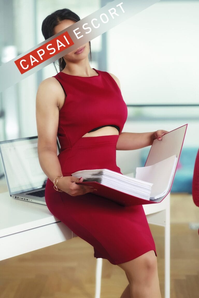 Escort Dame Katja in einem roten Kleid auf einer Tisch sitzend und einem Ordner in den Händen.