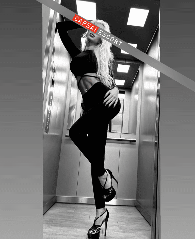 Escort Dame Aurelia in einem schwarzen kurzen Oberteil und Leggings mit schwarzen High Heels im Fahrstuhl stehend.