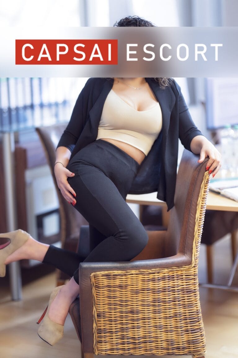 Escort Dame Liliana in einem beigen Top mit einem schwarzen Blazer und schwarzer Leggings mit beigen High Heels in kniender Pose auf einem Stuhl.