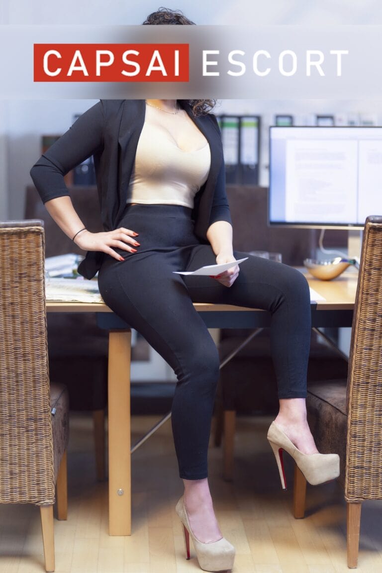 Escort Dame Liliana in einem beigen Top mit einem schwarzen Blazer und schwarzer Leggings mit beigen High Heels auf einem Tisch sitzend.