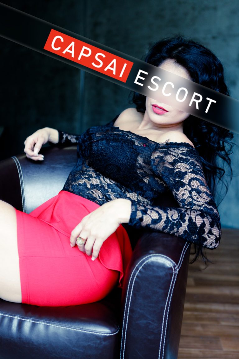 Escort Dame Luana in einem schwarzen Spitzenoberteil mit einem roten Rock im Sessel sitzend.