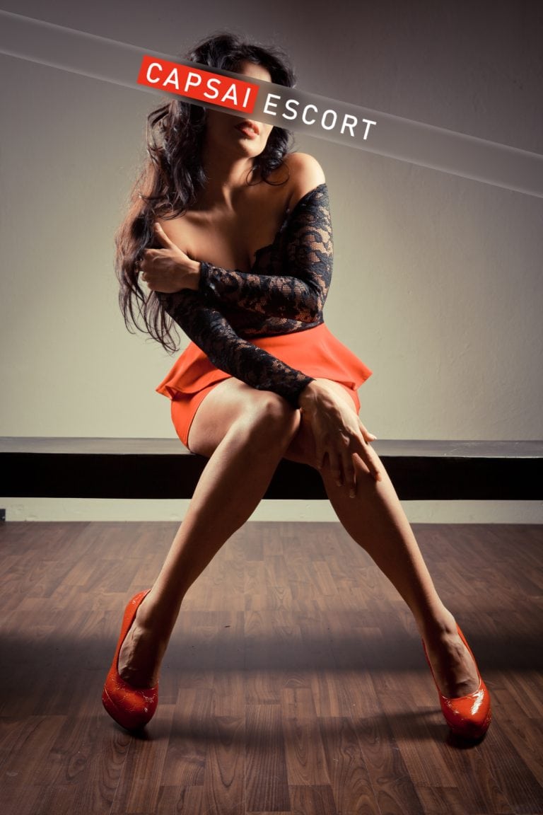 Escort Dame Luana in einem schwarzen Spitzenoberteil mit einem roten Rock und High Heels auf einer Bank sitzend.