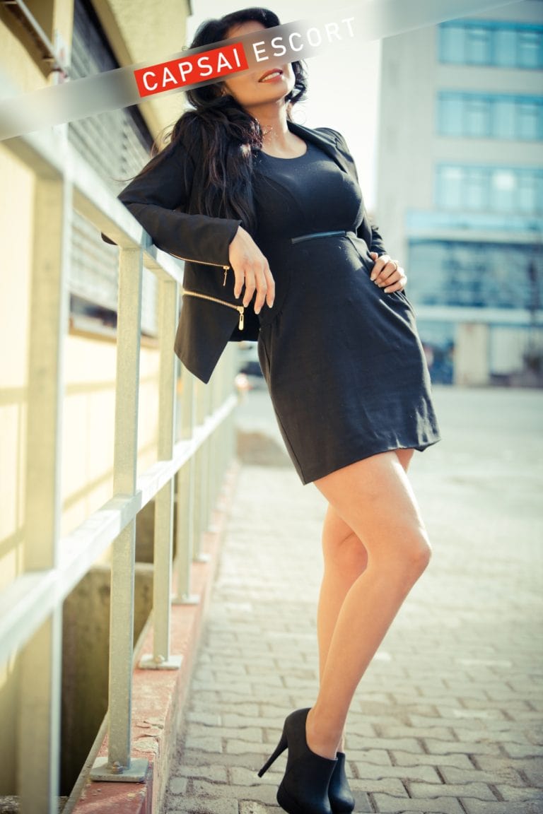 Escort Dame Luana in einem schwarzen Kleid mit High Heels und Blazer an einem Geländer gelehnt.