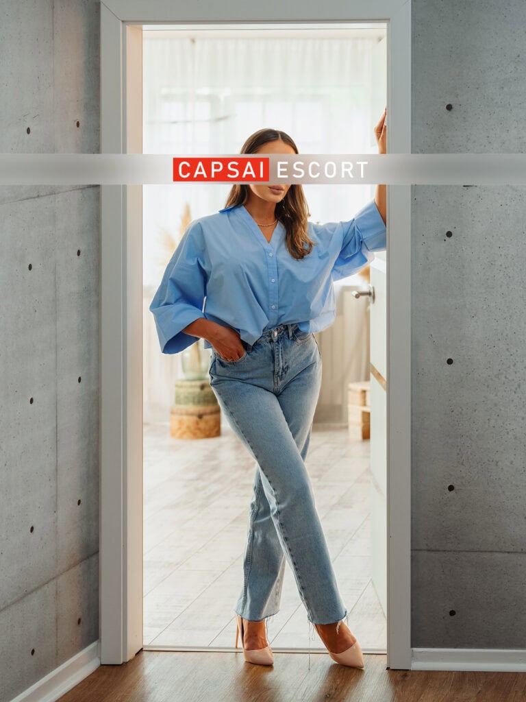 Escort Dame Mara in blauer Bluse und Jeans mit High Heels im Türrahmen stehend.