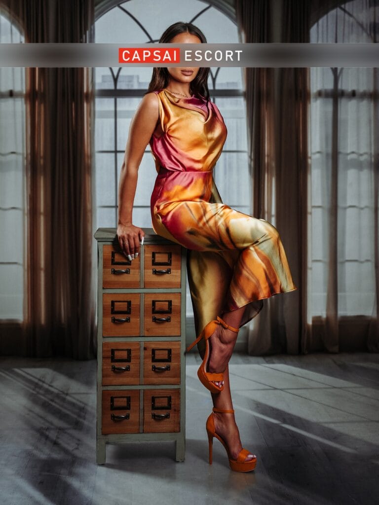 Escort Dame Mara in einem bunten langen Kleid mit orangenen High Heels an eine Kommode gelehnt.