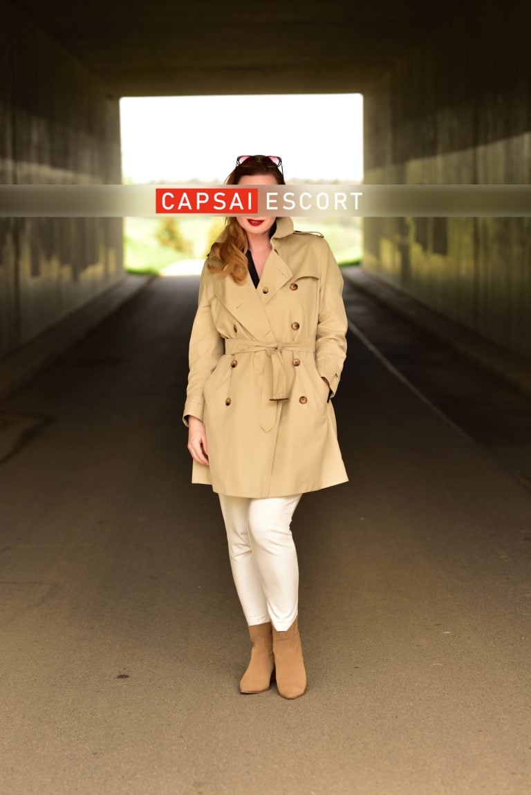 Escort Dame Marlene im Trenchcoat und weißer Hose draußen stehend.