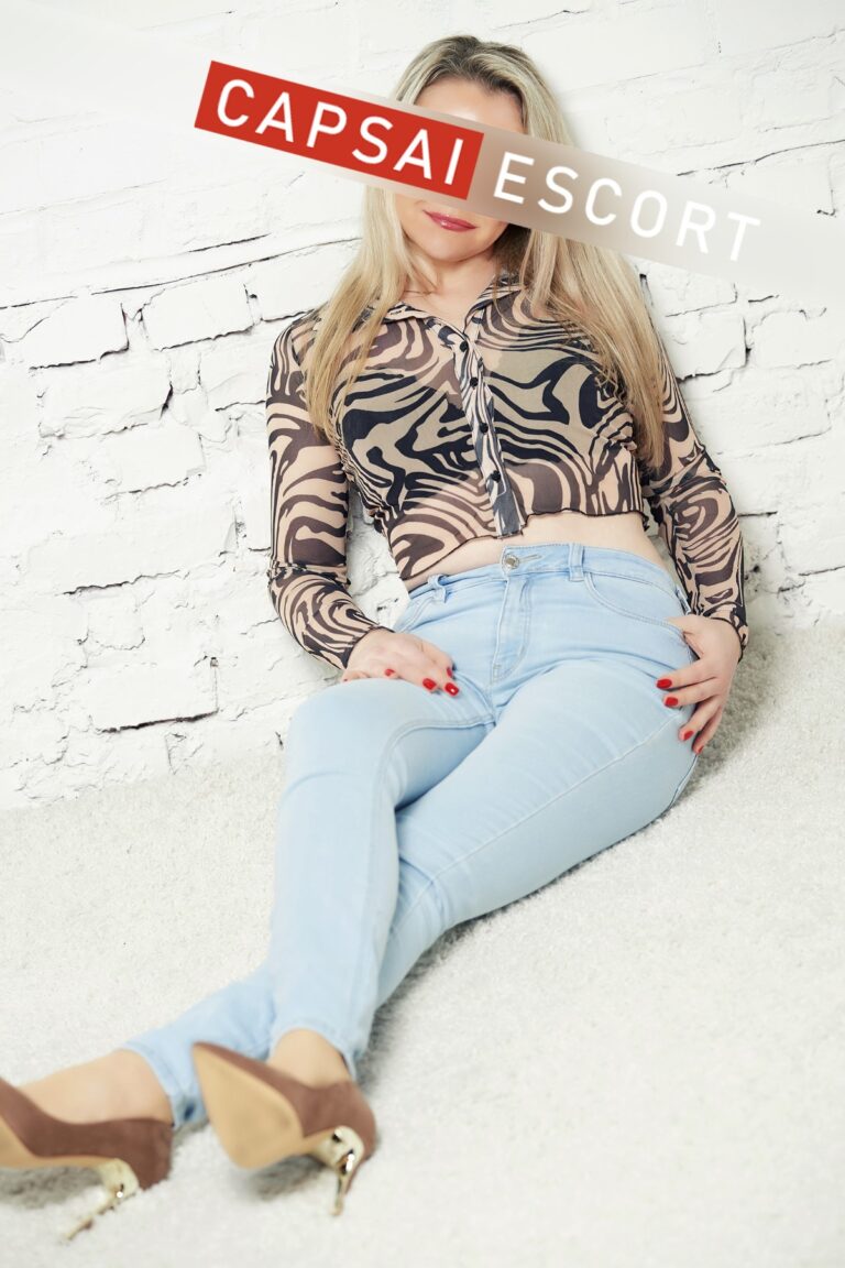 Escort Dame Melina in Jeans und transparenter Bluse und High Heels auf dem Boden sitzend.