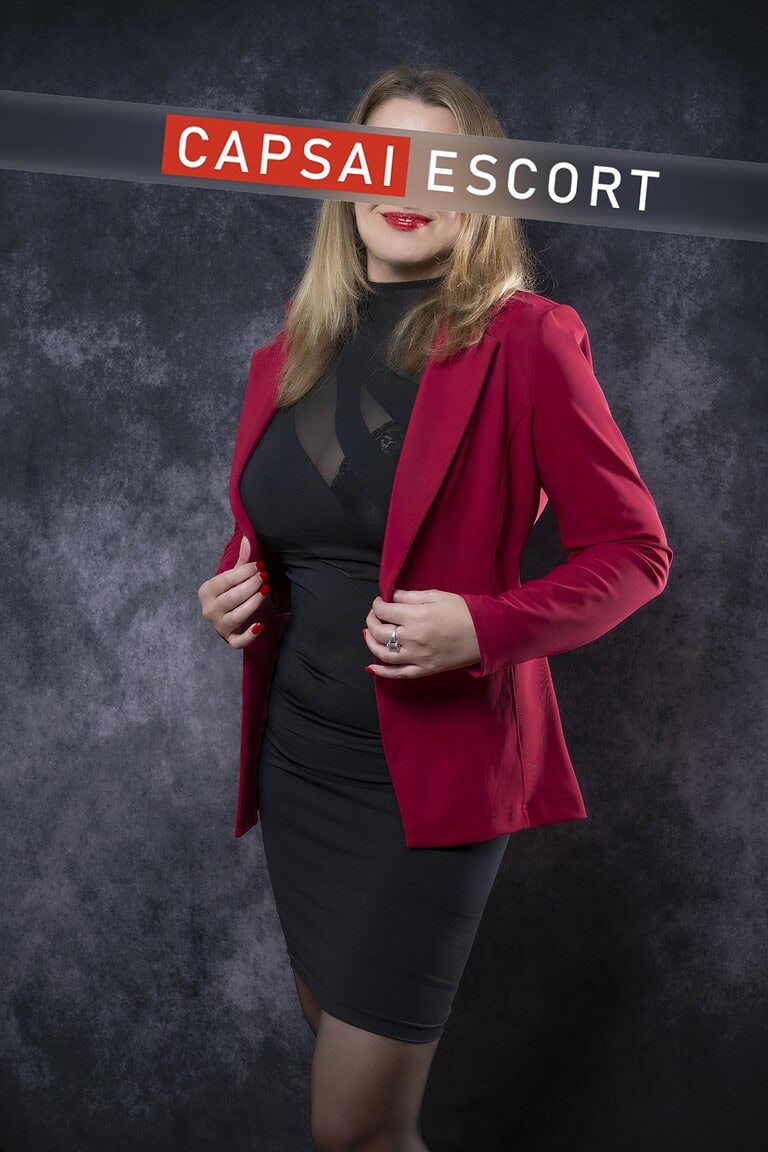 Escort Dame Michelle in einem schwarzen Kleid mit Nylons und einem roten Blazer.