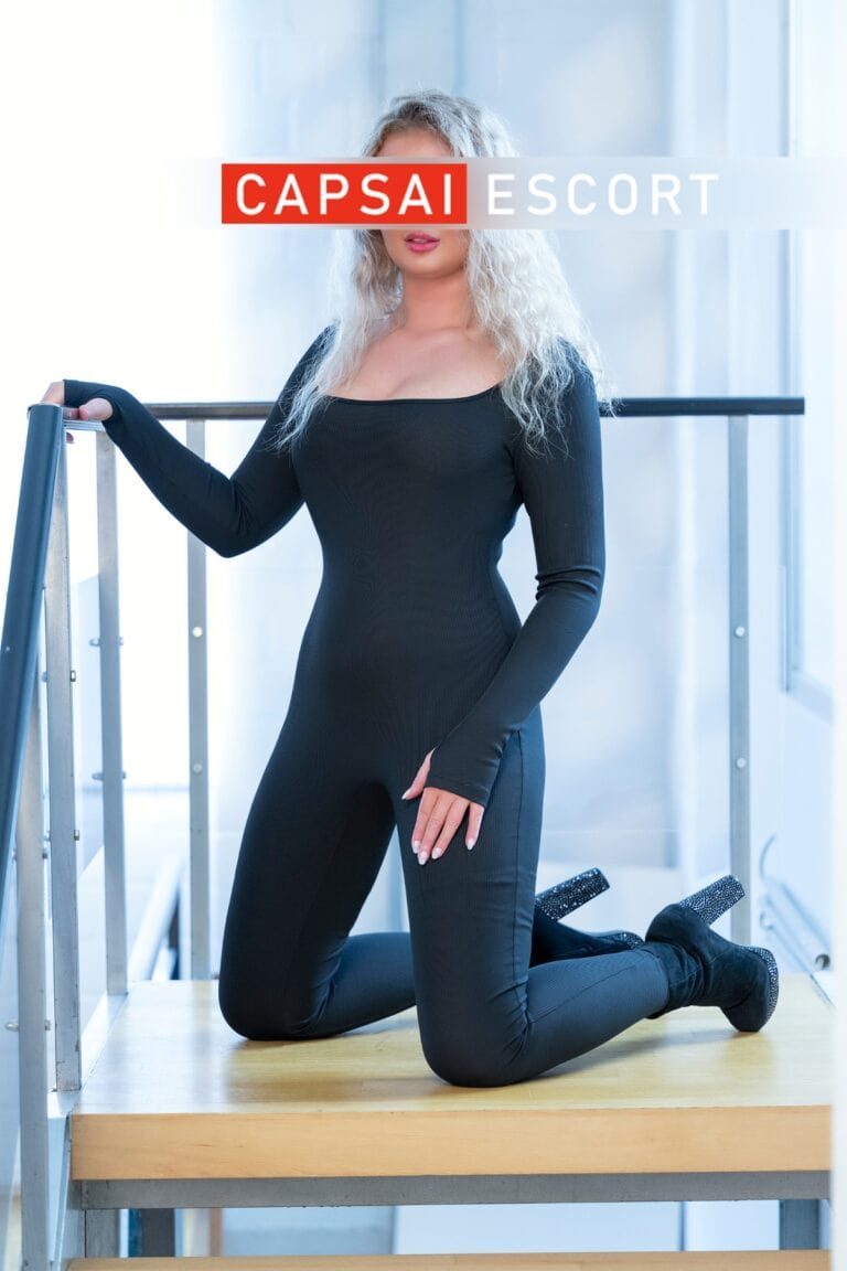 Escort Dame Leoni in einem schwarzen engem Overall und schwarzen High Heels in kniender Pose.