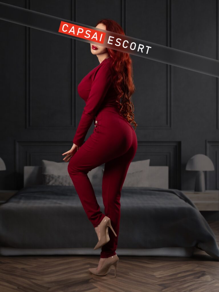 Escort Dame Nancy in einem rotem Jumpsuit und beigen High Heels in einer Pose mit Hinteransicht.