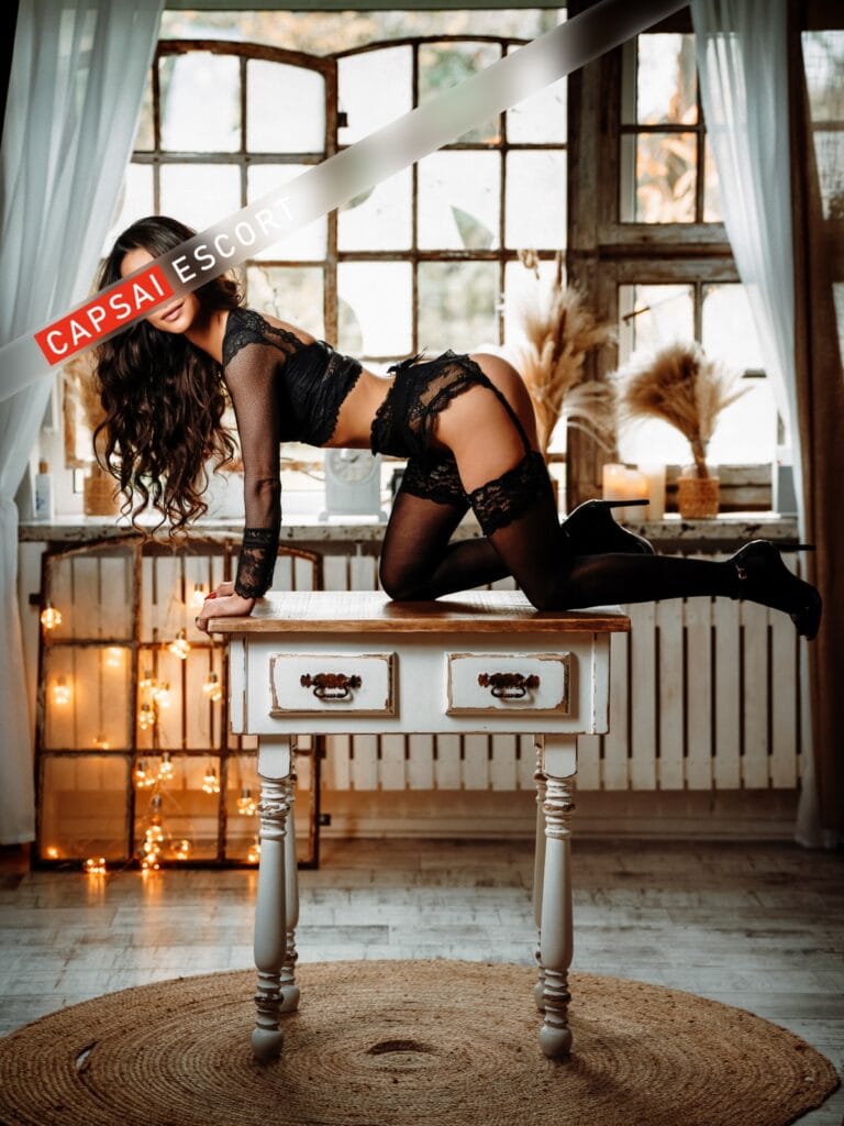 Escort Dame Nele in schwarzen Spitzen Dessous und schwarzen Nylons mit High Heels in knieder Pose auf einem Tisch, mit Ansicht von der Seite.
