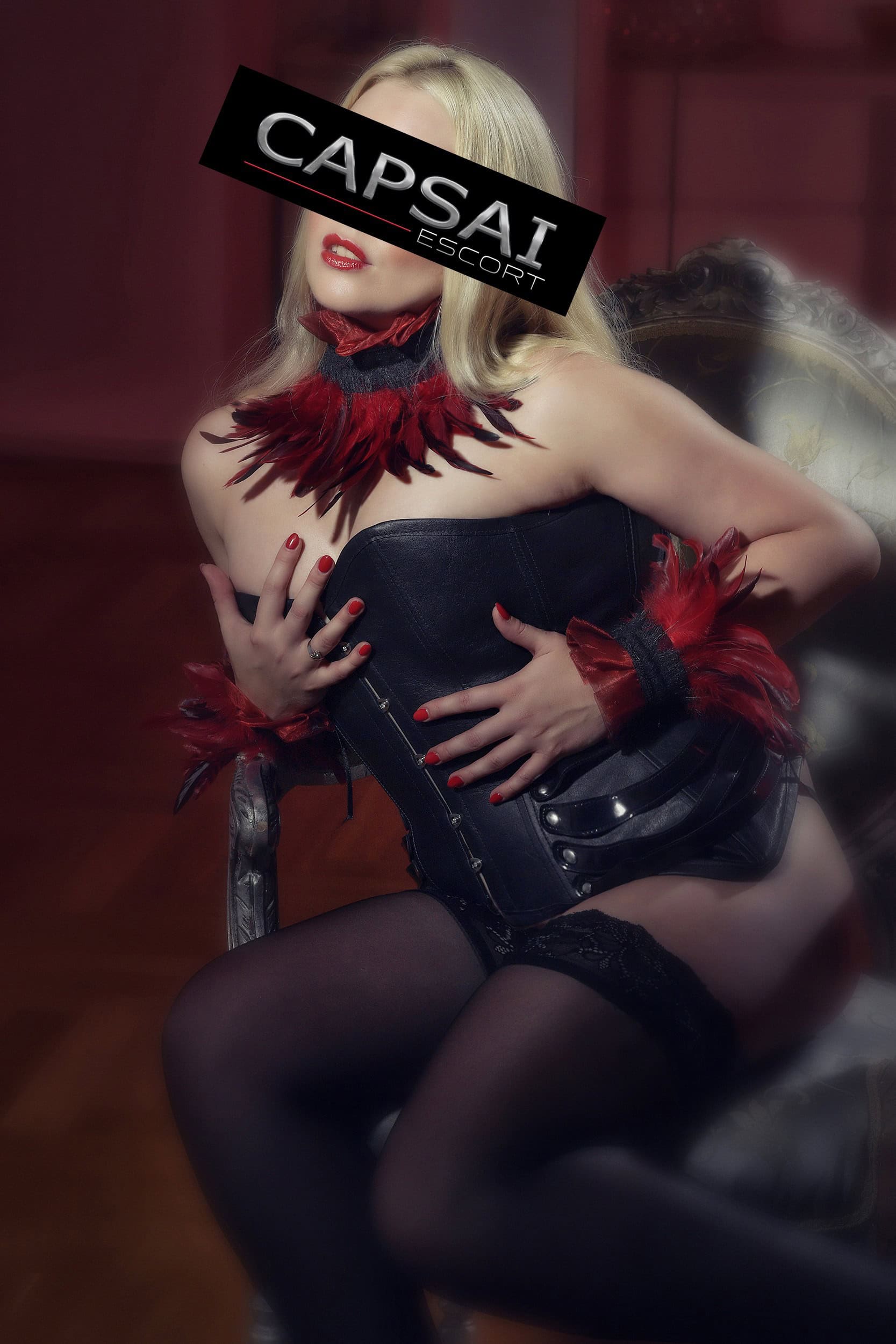 Escort Dame Estella in einer schwarzen Corsage mit schwarzem String und Nylons, um Hals und Handgelenke trägt sie Feder-Accessoires.