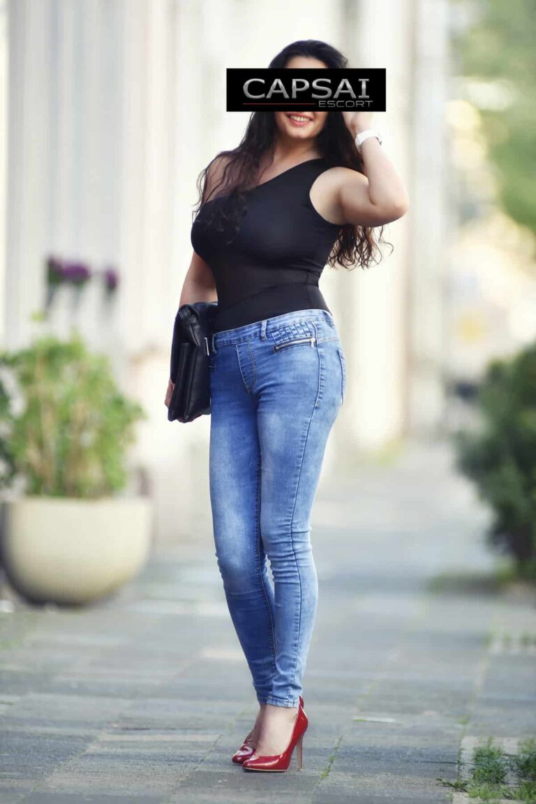 Escort Dame Kate in einem schwarzen Top und Jeanshose und roten Pumps draußen.